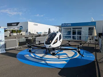 Un EH216-S blanc, cabine au centre et bras à hélices, posé sur une plateforme bleue devant le stand EHang.